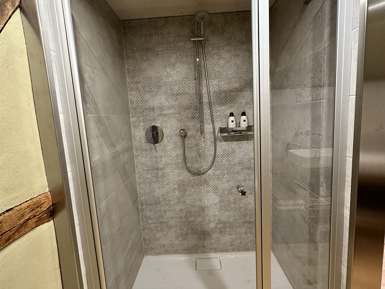 Geräumige Dusche mit Glastür, grau gemusterten Fliesen und Regendusche, seitlich das alte Fachwerk