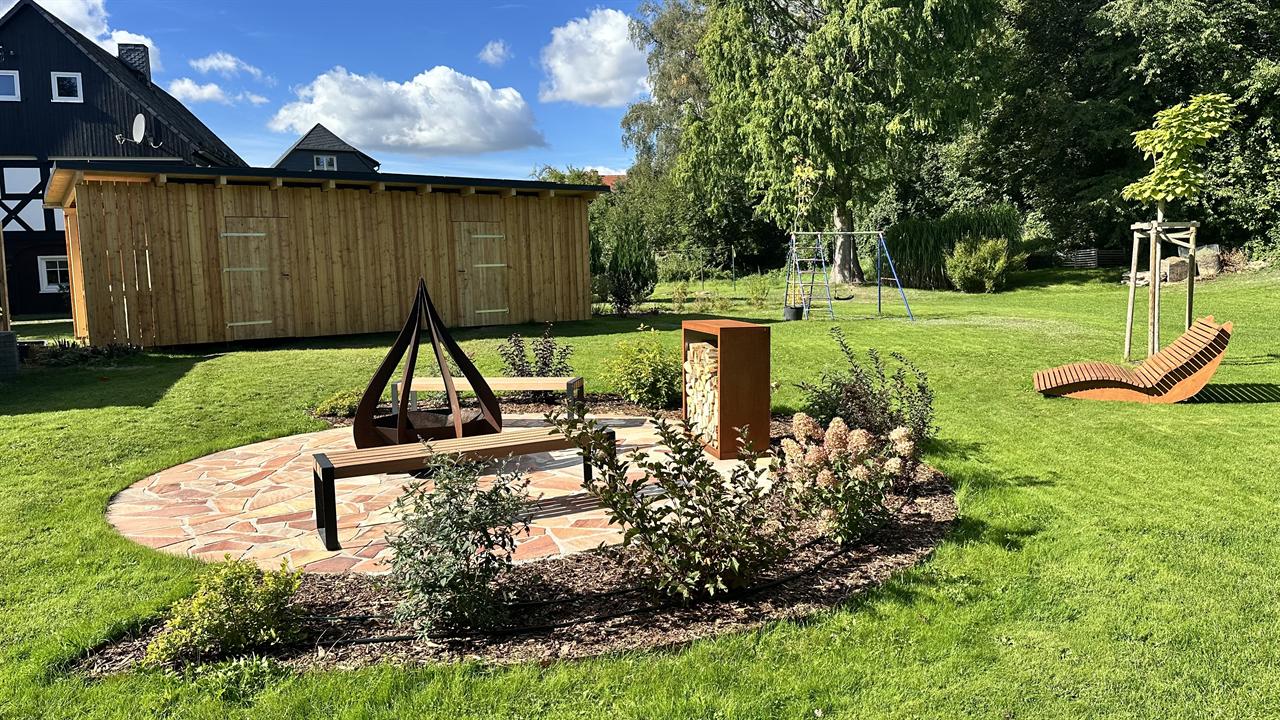Runder Natursteinplatz im Rasen mit Feuerschale aus Cortenstahl, zwei Holzbänken und gefülltem Holzlager, umringt von Hortensien; dahinter der große Holzschuppen und der weite Garten
