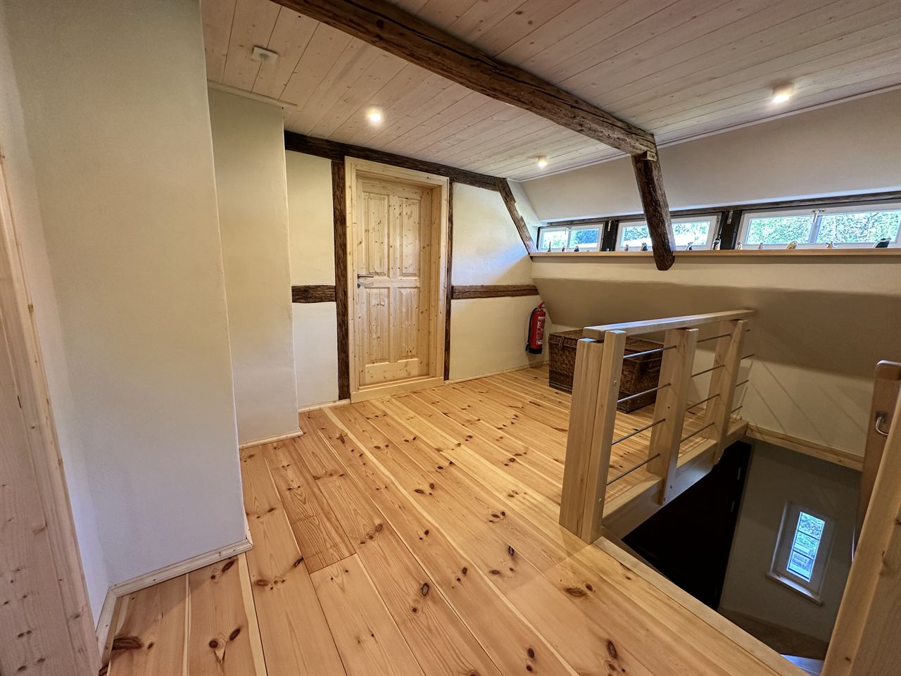 Flur im Obergeschoss mit hellem Dielenboden, altem Deckenbalken, einer Reihe kleiner Dachfenster und dem Holzgeländer über der Treppe