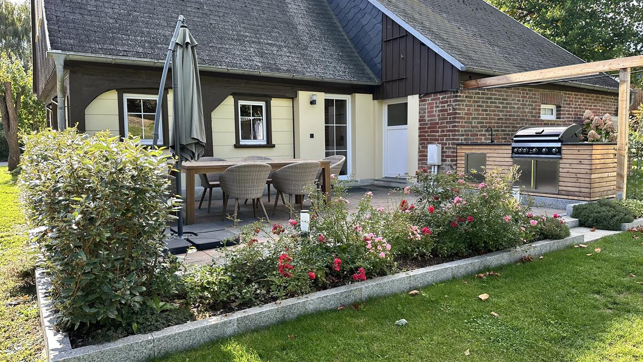 Gepflasterte Terrasse mit langem Holztisch, sechs Korbstühlen und Sonnenschirm unter dem Vordach; rechts an der Klinkerwand eine Außenküche mit eingebautem Gasgrill, davor ein blühendes Rosenbeet